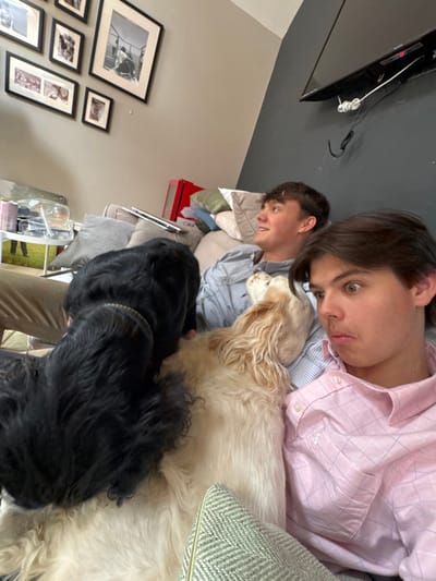 The image shows two young men and two dogs sitting on a couch. The couch is in a living room with a gray wall and pictures on the wall behind them. The man on the left is wearing khaki pants and a blue shirt. The man on the right is wearing a pink shirt. One dog, black, is partially covering the man on the left and the other, white, is partially covering the man on the right. The man on the right is looking toward the camera, while the man on the left is looking to the side. The image was taken in Sand Hutton, United Kingdom at the coordinates (latitude: 54.025, longitude: -0.933).