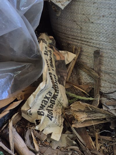 The image shows a pile of debris, including a crumpled newspaper with Cyrillic text, pieces of wood, and a transparent plastic sheet. The newspaper appears to be old and weathered. The debris is situated in an outdoor environment, possibly a back alley or a construction site. The location is identified as Sadina, Bulgaria. There are no people visible in the image. The scene is still and appears to be a snapshot of an abandoned or neglected area.