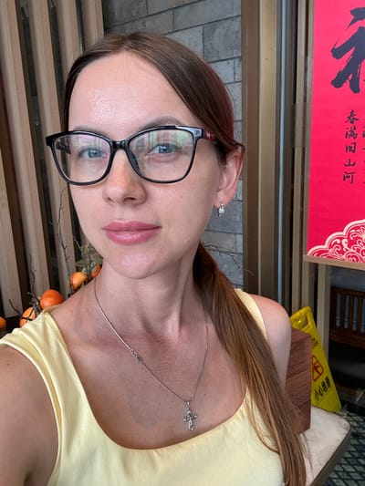 A young woman, appearing to be in her 20s or 30s, is captured in a close-up, angled selfie, looking directly at the camera with a neutral expression. Her long, straight brown hair is parted on the side, and she wears black-framed glasses that reflect her eyes. She has light pink lipstick and clear skin.

She is dressed in a light yellow top or dress. A delicate silver chain necklace with a small, intricate cross-like pendant rests on her collarbone. A small, flower-shaped earring or helix piercing adorns her right ear. Below her collarbone, a decorative branch adorned with small, round, orange fruits or berries is visible, suggesting seasonal decor.

The background indicates an indoor or sheltered outdoor environment, consistent with Fangshan District, China. To the top left, a vibrant red sign featuring bold black Chinese calligraphy is partially visible. Behind her, wooden slat-like structures are present. In the upper right background, a bright yellow "Wet Floor" caution sign with Chinese text stands. The ground beneath the structures appears to be paved with light-colored bricks or stones.

The visible text includes Chinese calligraphy on the red sign and "小小心地滑" (Xiǎo xīn dì huá), meaning "Caution: Wet Floor," on the yellow sign. The overall ambiance is calm and brightly lit, suggesting daytime.