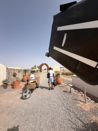 In Agafay, Morocco, a person walks away from the camera on a gravel path. The person is wearing a white t-shirt and blue shorts, carrying a brown bag over their left shoulder and a black bag in their right hand. To the left of the path, there are tents, wooden fences, and large terracotta pots with plants. A small, three-wheeled vehicle is parked on the left side of the path. In the background, an arched structure with an "M" logo is visible, with a clear blue sky above. A large, dark, curved object dominates the right side of the foreground.