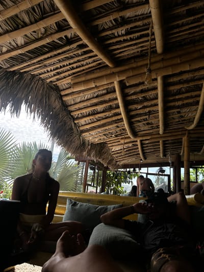 The image shows an outdoor seating area in Montañita, Ecuador, located at coordinates -1.825, -80.754. The seating area is covered by a large thatched roof supported by a framework of bamboo poles. A young woman in a bikini top and shorts is sitting on a bench on the left side of the image, facing towards the right. She is looking off to the right, and her face is partly obscured by a shadow. There is a laptop on the bench in front of her. A man in a black shirt and shorts is lying on a bench in the foreground, facing the left. He is obscured by a shadow and only his feet and part of his face are visible. A second man in a red and white patterned bathing suit and a dark shirt is sitting on a bench further back in the scene, facing towards the right. He is out of focus. The setting appears to be an outdoor restaurant or bar with a bamboo roof and benches for seating. There is a lot of greenery in the background. The overall mood of the image is relaxed and casual.
