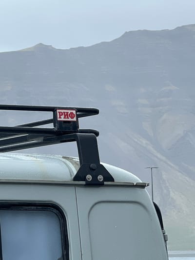 The image shows a close-up of the roof rack of a white van in Grundarfjörður, Iceland. The roof rack is made of black metal and has a red sticker on it that says "РИФ" in Cyrillic script. The background features a mountain range, visible through a hazy atmosphere. There is a single, tall streetlight in the distance. The image does not show any people.