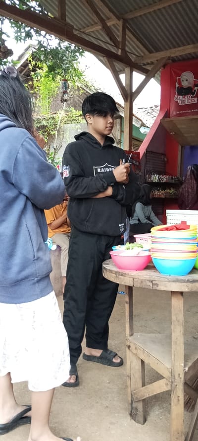 The image shows a scene of a young man standing outside of a small store in Cimaung, Indonesia, located at coordinates -7.074 latitude and 107.547 longitude. The man, wearing a black hooded sweatshirt with the words "Raiders" printed on it, is holding a cell phone in his right hand and appears to be looking at it.  His left arm is folded across his chest. He is standing near a wooden table on which there are a variety of colorful plastic containers stacked on top of each other. There are a few other people in the background, but they are blurry and their actions are not clear. Behind the table, a sign is visible on the side of a building, which appears to be advertising "SBLAK TEH DEWI" (translated to "Tea Dew"). The ground is dirt and there are some plants visible in the background. The overall tone of the image is casual and mundane.