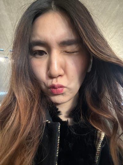 A woman winks and purses her lips, posing for a selfie. Her long, wavy hair transitions from dark brown at the roots to a lighter, reddish-brown towards the ends. She is wearing a dark jacket with a textured or possibly furry collar. Her skin is fair with a natural complexion. The background is blurred and indistinct, likely an interior space.