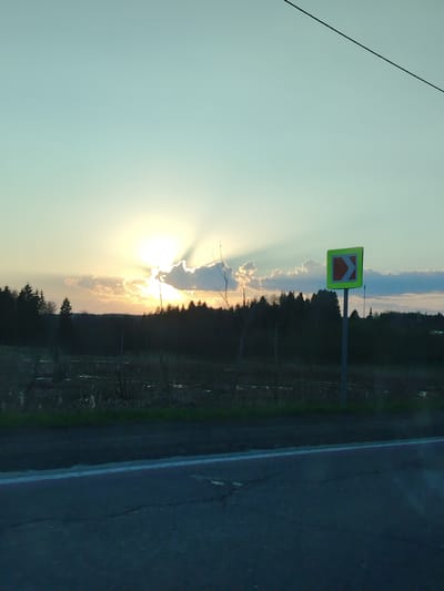 The image shows a rural roadside scene in Malyi Dubovik, Russia. The sun, partially obscured by clouds, casts crepuscular rays across the horizon. A right-pointing road sign is visible on a metal pole. Behind the sign, there is a dark line of trees along the horizon. In front of the trees is a field. The foreground features a road with a white line. A powerline is visible in the upper right corner of the image.