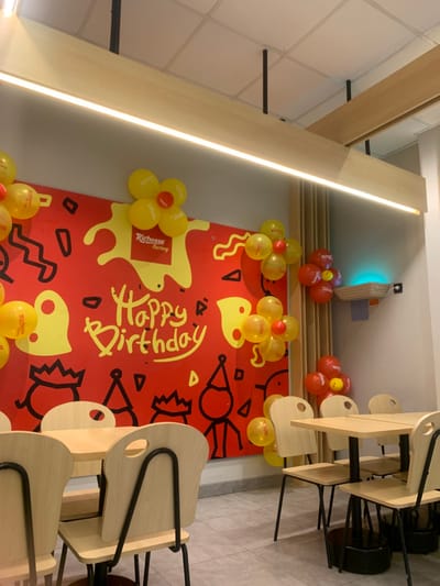 The scene depicts the interior of a Richeese Factory restaurant located in Calukan RT 1 RW 5 Keboansikep, Indonesia. The area is set up for a birthday celebration. The focal point is a large red banner covering a wall, featuring yellow abstract shapes, black line drawings of party hats and stick figures, and the prominent yellow text "Happy Birthday." The "Richeese Factory" logo is visible on the banner. Numerous yellow and red balloons, many bearing the "Richeese Factory" logo, are affixed to the banner and adjacent walls, some arranged in flower-like clusters.
In the foreground and midground, multiple light-colored wooden tables with dark metal pedestal bases are arranged. Around these tables are matching light-colored wooden chairs with dark metal frames, each featuring an oval-shaped cutout in the backrest. No individuals are present in the image.
The ceiling consists of white grid panels, from which several linear LED light fixtures extend, providing illumination. Exposed black pipes or conduits are visible against the ceiling. To the right of the main banner, a section of the wall features vertical light-toned wooden slats, above which a wall-mounted fixture emits a distinct blue light. The floor is covered with grey tiles. The overall environment is brightly lit and appears clean and organized.