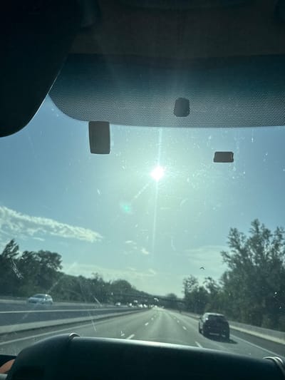 The image, taken from inside a vehicle, captures a view through the windshield on a sunny day. The sun is bright and positioned high in the sky, creating glare and lens flare. There are several small, dark rectangular objects adhered to the windshield. The road ahead is a multi-lane highway with cars visible in the distance. Green trees and foliage line the sides of the highway. The overall environment suggests a drive through a rural or semi-rural area. There are no identifiable landmarks specific to Avignonet-Lauragais, France in the image.