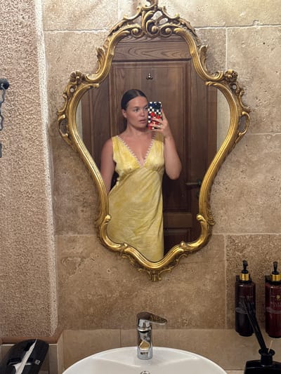 A woman is taking a mirror selfie in a bathroom. She is wearing a yellow dress with a textured pattern and white trim around the neckline. She is holding a smartphone with a red, white, and blue checkered case in her right hand, which is raised to take the photo. Her hair is dark and pulled back. The mirror is ornate with a gold-colored, carved frame. The background reflected in the mirror appears to be a wooden door. The bathroom walls are covered in beige stone tiles, and a textured beige stone ledge runs along the bottom of the wall. To the right of the mirror, a white sink with a chrome faucet is visible, and next to it, a black dispenser with white paper products. Above the mirror, mounted on the wall, are two dark brown dispensers with gold accents. The lighting suggests it is daytime. The overall ambiance is one of a quaint, perhaps historic, establishment. The scene is set in Stanišići, Montenegro.