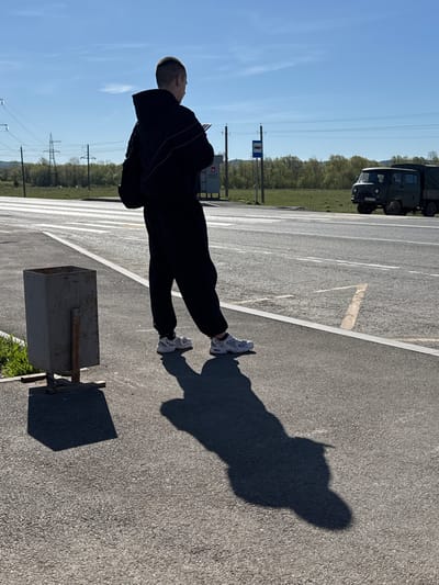 The image shows a person standing on an asphalt surface, likely a parking lot or roadside, in Krilovo, Russia. The person is wearing a dark tracksuit and white sneakers. Their shadow is cast on the asphalt beside them. In the background, there is a military truck, a bus stop sign, electrical pylons, trees, and a blue sky. A metal trash receptacle is also present. The person appears to be looking upwards.
