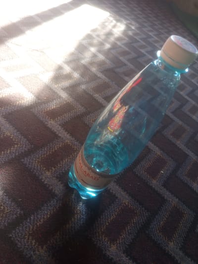 A partially filled, translucent blue plastic water bottle with a light-colored cap rests diagonally on a textured, dark brown and blue patterned fabric surface. Sunlight streaks across the fabric from the upper left corner. A label is visible on the bottle, with text that includes "JALAL-ABADI" and "PREMIUM MINERAL WATERS".