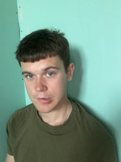 The image shows a head and shoulders shot of a young white male with short, dark brown hair and blue eyes. He is wearing a dark olive green t-shirt and a silver chain necklace. He is standing in front of a teal-colored wall. The lighting suggests an indoor setting, potentially a room under construction or renovation, given the presence of what appears to be drywall and some light fixtures or imperfections on the wall. The individual is looking directly at the camera with a neutral expression. There are no other individuals or objects of significance visible in the frame. The location of Аснежыцкі сельскі Савет, Belarus is not depicted or implied by any visual cues in the image.