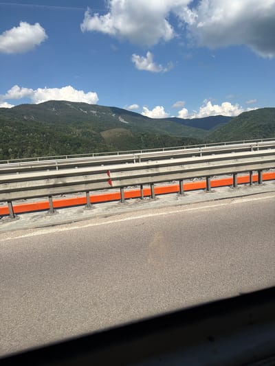 The image shows a view from a bridge with a mountainous landscape in the background. The bridge has a metal guardrail with orange accents. The sky is blue with white clouds. The mountains are covered in green trees, and there are some bare patches on the slopes. The foreground shows the asphalt road of the bridge. No people are visible in the image. This scene is consistent with the location of the Vrachanski Pass bridge, which is located near the town of Vrachesh, Bulgaria, and offers views of the Balkan Mountains.