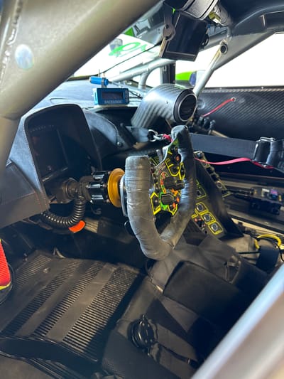 The image captures the interior of what appears to be a race car, likely at the Vallelunga Circuit located near Campagnano di Roma, Italy. The focus is on the steering wheel, which is a specialized racing wheel with numerous controls and wrapped in tape. The steering column is visible below the wheel. The surrounding interior is sparse, with carbon fiber paneling and racing harnesses. There's a small digital display mounted on the roll cage, along with other electronic components. The overall impression is that of a cockpit optimized for performance driving.