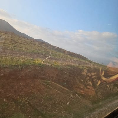 The image displays an outdoor landscape in Ouahat Sidi Brahim, Morocco, viewed through a reflective transparent surface. The scene is dominated by a gradual upward-sloping hillside composed of brown earth with interspersed patches of green vegetation and yellow flowering plants. A rudimentary fence line, constructed from several vertical posts and horizontal wires, runs across the mid-section of the slope. Beyond this fence, the terrain continues to rise towards a distant, darker mountain range visible on the left side of the frame. The sky above is partially clear blue with scattered white clouds. A prominent reflection on the right side of the transparent surface reveals a partial human face, specifically an eye, eyebrow, and forehead, alongside what appears to be colorful fabric in red and yellow/gold tones. No individuals are directly visible within the landscape itself.
