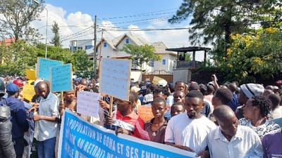 The scene depicts a dense crowd of individuals engaged in an outdoor protest or demonstration in the city of Kabinzi, Rwanda. The foreground features numerous people, predominantly adults and some youth, standing closely together. Several individuals hold handwritten signs of various colors, including light blue, white, and yellow, displaying text in what appears to be a local language. A large blue and white banner, partially visible, stretches across the lower portion of the image, with French text reading "POPULATION DE GOMA ET SES ENVIRO[NS]" and "DU RWK AU RETRAIT DE..." suggesting a focus on the population of Goma and withdrawal related to Rwanda. On the far left, at least two individuals in dark blue uniforms, helmets, and protective gear, consistent with law enforcement, are visible. The crowd is diverse in age and attire, with many looking towards the front of the gathering.

In the background, under a partly cloudy blue sky, multiple two-story buildings with light-colored facades, pitched gray roofs, and blue-framed windows are visible. Lush green trees and shrubs, some bearing yellow flowers, line the street and properties. A dark wooden structure, possibly a guard post or observation deck, stands above a wall on the right side of the image. Overhead utility lines are present across the sky. The overall environment suggests an urban or semi-urban setting during daylight hours.