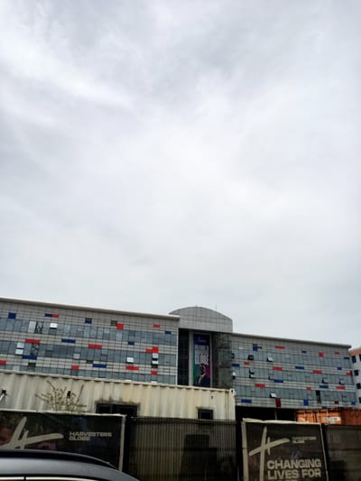 The image displays the exterior of a modern, multi-story building under an overcast sky, set in what is identified as Eputu Town, Nigeria. The building features a distinctive facade composed of numerous gray panels and windows, accented with scattered red and blue rectangular elements. Several windows on the building are partially open.

A large, vertical banner hangs on the left side of the building, displaying the text "WELCOME TO HARVESTERS GLOBE" and featuring a portrait of a man in a dark suit. In front of the main building, several shipping containers are visible; some are off-white/beige, and at least one is a rusty orange-brown. One container bears the identification "SJBU 151984 7".

A dark, chain-link fence runs across the mid-ground, separating the viewer from the containers and building. Behind this fence, two large signs are present. The left sign is black with white text reading "HARVESTERS GLOBE" and partially visible "CHANGING LIVES FOR". The top of a dark vehicle is visible in the bottom left corner of the frame, suggesting the photo was taken from a car or similar vantage point. The sky is entirely covered with light gray clouds, indicating an overcast or cloudy daytime. No specific activity or people are clearly depicted, presenting a static view of the building and its immediate surroundings.