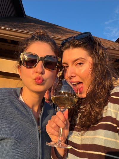 Two women are taking a selfie outdoors in front of a house with a brown shingled roof. The woman on the left is wearing a blue zip-up sweater and tortoise-shell sunglasses, with her hair in a bun. She is puckering her lips. The woman on the right is wearing a striped sweater and black sunglasses perched on her head. She is drinking from a wine glass, with her tongue extended. There is a man visible in the background between the two women. The sky is clear and blue, and the setting sun casts a warm glow on the scene. The location is Mont-Tremblant, Canada.