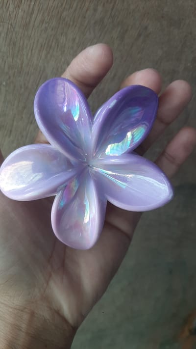 A person's hand holds a purple, iridescent, five-petal flower ornament against a wooden background. The ornament is centered in the frame, with the hand supporting it from below and the side. The wood grain is visible and runs diagonally from the top left to the bottom right. The hand shows visible lines and texture. The ornament has a glossy finish and reflects light in various colors, including blue, green, pink, and yellow, creating a shimmering effect. The background is out of focus, emphasizing the hand and the ornament. The image is taken from a top-down perspective.