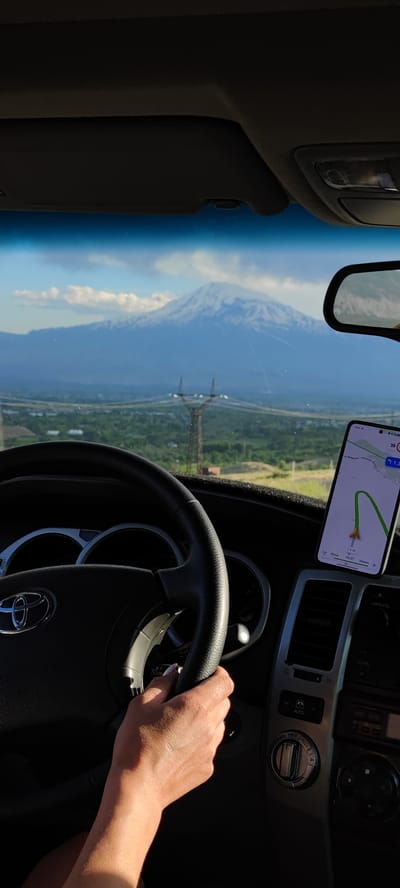 The image shows a view from the driver's seat of a car, looking through the windshield at a large, snow-capped mountain in the distance. The sky is partly cloudy. In the foreground, the steering wheel and dashboard are visible, with a smartphone mounted on the dashboard displaying a navigation app. A person's hand is on the steering wheel. The landscape outside the car appears to be rural, with green fields and trees. Power lines and a transmission tower are also visible between the car and the mountain. The sun is casting shadows within the car, indicating it is likely daytime. This scene is consistent with a view of Mount Ararat from the vicinity of Lanjazat, Armenia, given the presence of the mountain and the type of landscape.