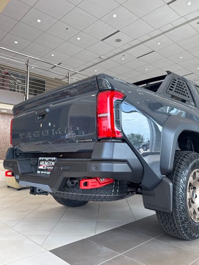 The image shows the rear end of a gray Toyota Tacoma truck parked inside a dealership in Palatine, Illinois. The dealership appears to be Arlington Toyota, evidenced by the license plate frame that says "Arlington Toyota Palatine" and includes the website "TOYOTAARLINGTON.COM" along with a phone number. The truck has a spare tire mounted on the back and a red ARB recovery point visible on the bumper. The "TACOMA" model name is embossed on the tailgate, which also features "i-FORCE MAX" branding. The backdrop consists of a well-lit interior with tiled floors and modern architectural elements like a staircase and a patterned ceiling.