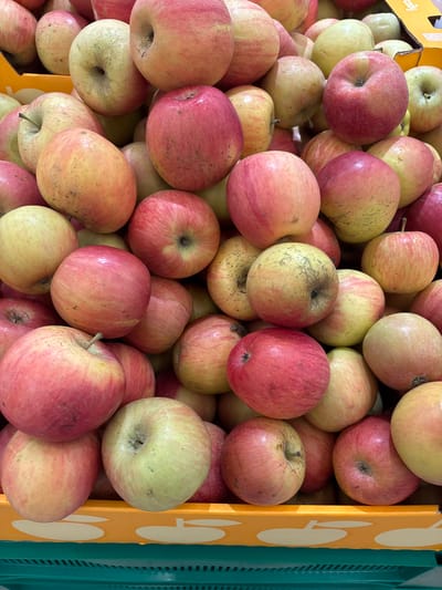 A close-up, eye-level shot shows a large pile of ripe, red and yellow apples filling the frame. The apples are various sizes and shades of red and yellow, with some showing streaks and blemishes. They are densely packed together, with some apples resting on top of others, creating a textured surface. The background is blurred, but appears to be a market or grocery store setting, with hints of orange and green packaging visible at the bottom of the frame. The lighting is bright and natural, highlighting the vibrant colors of the fruit. No people are visible in the image. The scene is consistent with a display of produce in a commercial setting in Kuzmolovsky, Russia.
