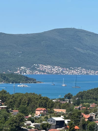 The image captures a panoramic view of a coastal landscape, likely in Kavač, Montenegro, on a clear day with a bright blue sky. The foreground features a dense expanse of green trees and several residential buildings with terracotta roofs, nestled on a hillside. These structures vary in size and architectural style, suggesting a mix of older and more modern dwellings.

Further back, a serene body of water, possibly a bay or gulf, is visible. Several boats, including sailboats and a large white yacht, are scattered across the water, indicating recreational activity. Along the shoreline of the water, a town or village with numerous white and red-roofed houses is densely built on the slopes of a large, tree-covered mountain that dominates the background. The mountain itself has a rugged, natural appearance, with some telecommunication towers sparsely visible on its summit.

The overall scene depicts a tranquil coastal environment where nature and human habitation coexist, characterized by lush greenery, clear waters, and a picturesque settlement against the backdrop of a majestic mountain.