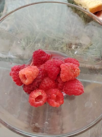 A close-up, overhead shot shows a collection of raspberries inside a clear glass bowl. The raspberries are a vibrant red, with one slightly lighter, more orange-hued berry in the center of the cluster. The bowl is empty except for the fruit, and the background is blurred, showing hints of greenery and what appears to be a slice of bread or toast in the upper right corner of the image. The lighting suggests it might be daytime. There is no visible text. The scene appears to be indoors, possibly in a kitchen or dining area, given the presence of the food items.