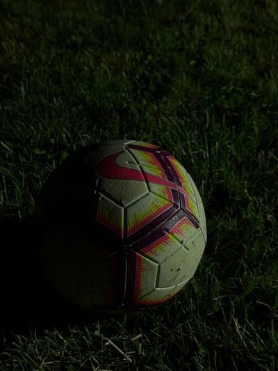A soccer ball rests on green grass. The ball is partially illuminated by a light source, casting a shadow to the left. The grass appears to be a lawn or a field. The image does not contain any elements that would indicate a location in the city of Taban, Bulgaria.