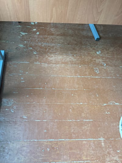 The image depicts a close-up, overhead view of a worn wooden floor. The floorboards are oriented horizontally and appear to be made of a light-colored wood, stained a reddish-brown. The surface is heavily scuffed and scratched, with areas where the paint or finish has chipped away, revealing lighter wood underneath. There are distinct horizontal lines running across the image, likely representing the gaps between the floorboards or ingrained scratches. In the upper left corner, a portion of a light blue object, possibly furniture or a piece of equipment, is visible. In the upper center, a silver-colored, L-shaped metal object is partially visible, resting on the floor and extending upwards. The background above the floor appears to be a wall made of similar light-colored wood paneling. The scene suggests an interior space that has seen significant use.