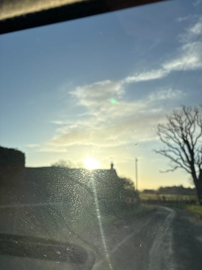 The image presents a rural landscape in Ruyton-XI-Towns, United Kingdom, captured through what appears to be a vehicle's front window. The window surface is notably dirty, covered with a fine layer of dust, small particles, and streaks that interact with light, causing significant glare and lens flare effects, particularly from the prominent sun. A dark, blurred element, consistent with a vehicle's interior trim or dashboard, occupies the top edge of the frame.
The environment outside is dominated by a bright, low-angle sun positioned towards the central-right, suggesting either sunrise or sunset. The sky above is clear to partly cloudy, displaying a light blue hue with scattered white clouds. Silhouetted against the bright horizon are several elements: on the left-center, the dark outline of a building, likely a house, is visible with a discernible roofline and chimney. To the right, a bare, deciduous tree stands with its intricate branch structure. A thin, vertical structure, possibly a utility pole, is faintly discernible between the building and the tree. In the foreground, a dirt or gravel path or road curves from the lower-center towards the right, leading into what appears to be open fields or green land. No individuals, animals, or specific actions are identifiable within the scene.