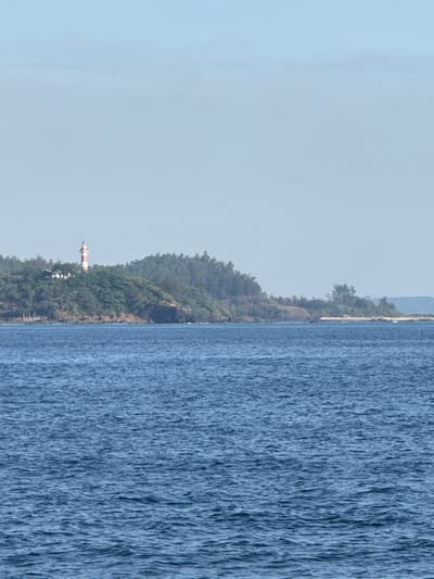 The image depicts a coastal scene with a body of blue water in the foreground and a verdant, hilly coastline in the background. A prominent lighthouse with red and white horizontal stripes stands on the highest point of the hill, accompanied by a white building. The coastline is covered in dense green foliage, with trees and shrubs extending down to the water's edge. The sky is a pale, hazy blue, suggesting either early morning or late afternoon light, or possibly a slightly overcast condition. The water exhibits ripples and small waves, indicating some movement. There are no people or animals visible in the image. The location is identified as An Hai, Vietnam, implying a maritime or island setting.