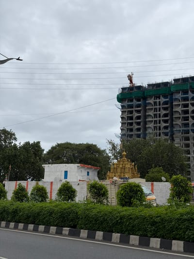 The image depicts a street scene in Shamshabad, India, on a cloudy day. In the foreground, a paved road is visible with a black and white striped curb and a row of lush green bushes lining the edge. Behind the bushes, a white concrete wall with red vertical stripes separates the street from a white building with an orange-tiled roof and blue windows. To the right of the building, a golden tiered structure, possibly a temple or shrine, stands behind a partially obscured white truck. Further in the background, a large, unfinished multi-story building under construction is prominently featured, with a red crane visible at the top. The sky is overcast with thick grey clouds. There are no people clearly visible in the image.
