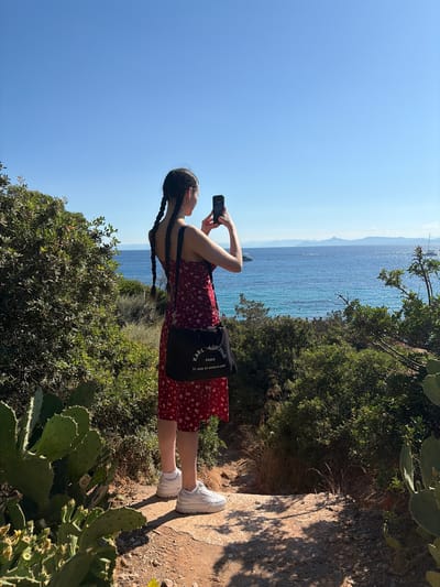 A young woman with braided hair stands on a dirt path overlooking the ocean, holding up a phone to take a picture. She is wearing a red floral dress and white sneakers, and has a black messenger bag over her shoulder. The ocean is a vibrant blue, with a few boats visible in the distance. The path is lined with green bushes and cactus plants. The sky is clear and blue. The image appears to have been taken in a coastal area, possibly a park or nature reserve. The specific location in Vouliagmeni Municipal Unit, Greece, is not discernible from the image itself, but the scene is consistent with a Mediterranean coastal landscape.