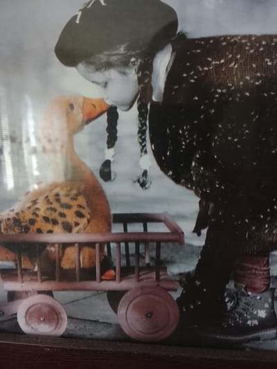 This is a black and white photograph of a young girl with braided hair wearing a dark beret and a patterned coat. She is leaning over to kiss a stuffed toy duck. The duck is in a small wooden wagon with red wheels. The girl appears to be holding the duck's beak with her lips. Her expression is one of affection and tenderness. The background is blurred and indistinct, making it difficult to determine the exact setting, but it appears to be indoors or in a sheltered outdoor area. The lighting suggests daytime. There is no visible text in the image.