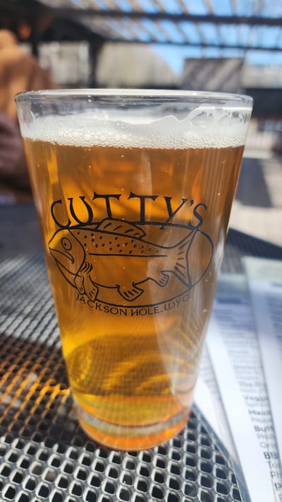 A glass of beer sits on a metal grate, with the words "CUTTY'S" and "JACKSON HOLE, WYO." printed on its side. The beer is light amber in color and has a foamy head. The background includes blurred images of people and objects. The overall setting suggests an outdoor dining or patio area. Given the inscription, this scene likely takes place in or around Jackson Hole, Wyoming, in the United States.