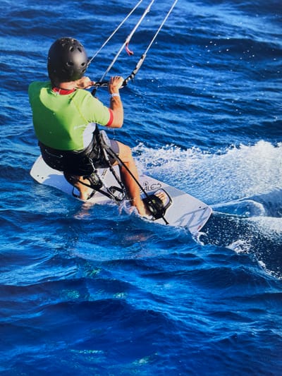 A man kiteboards on deep blue water, spraying a large wave to his right. He wears a black helmet, a lime-green and red short-sleeved shirt, dark shorts, and a black harness. He's gripping the kite's control bar with both hands, lines extending upwards from the bar. He is riding a white kiteboard with black bindings. The water is deep blue with subtle ripples and white spray. The location is Bacong, Philippines.