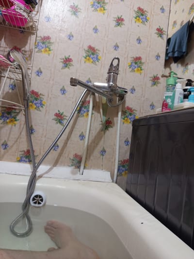 The image depicts a bathroom scene, likely in an apartment in Noyabrsk, Russia. The focal point is a bathtub filled with water, in which a person's foot is visible. A shower head and hose are positioned over the tub. The wallpaper has a floral pattern. To the right of the bathtub, a dark-colored washing machine is partially visible, topped with several bottles of toiletries. A blue towel hangs on the wall in the background.