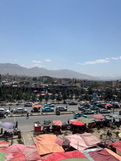 This is an elevated, wide-angle, eye-level, outdoor, daylight, vertical, full shot of a busy street scene in Kabul, Afghanistan, with a market area in the foreground and a city and mountainous background. The sky is a clear, light blue with a few scattered white clouds. The city, with a mix of modern and older buildings, stretches out in the midground, with a prominent mosque minaret visible to the left. In the background, hazy, arid mountains frame the cityscape.

The foreground is dominated by an outdoor market with numerous colorful tents and umbrellas providing shade. These range in color from various shades of red, pink, and orange to green and brown, some with intricate patterns. Stalls appear to be set up under these awnings, with goods and merchandise potentially displayed. A few individuals are visible within the market area, some appearing to be vendors or shoppers.

A wide, paved street runs horizontally across the midground, with several lanes of traffic. Numerous cars are present, including sedans, SUVs, and what appear to be taxis or similar passenger vehicles, in various colors such as white, silver, light blue, and dark gray. People are walking on the sidewalks along the street and crossing it. One person is walking on the sidewalk to the left of the street, while several others are visible in the street itself, including a person walking across the road in the center.

A line of trees with dark green foliage runs along the edge of the street, separating it from the market area. Some of the market stalls are set up behind this line of trees.

The overall impression is a vibrant, active urban environment on a sunny day, likely a marketplace and main thoroughfare in Kabul.