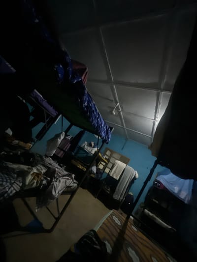 The image depicts a dimly lit dormitory room, likely in Ise-Ekiti, Nigeria, with multiple bunk beds. The room is cluttered with bedding, some appearing to be neatly made, while others are disheveled. Clothes are visible, some folded and stacked on shelves or furniture, while others are draped over the beds. The walls are painted a light blue, and the ceiling is a textured white with visible paneling and light fixtures. The overall impression is of a functional, perhaps crowded, living space.