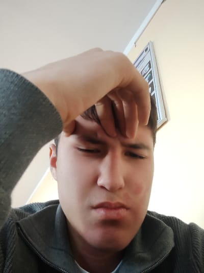 This image, captured in Rishtan, Uzbekistan, depicts a male individual in a close-up, upward-angled shot. The subject, with dark hair, has closed or tightly squinted eyes, a furrowed brow, and a slightly pursed mouth. A distinct reddish discoloration is present on his right cheek. His right hand is positioned with fingers resting against his forehead. He is attired in a dark grey, ribbed, zippered garment worn over a lighter undershirt, visible at the collar. The background consists of a light-toned wall. A white-framed object, containing indistinct graphic elements, is mounted on the wall in the upper right quadrant. A white ceiling is visible in the upper left portion of the frame. The scene is illuminated by bright, even light.