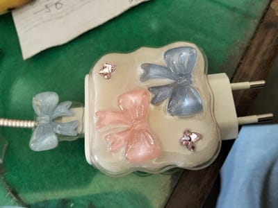 A decorative USB charger with two prongs is shown. The charger's face has a translucent, milky-white background adorned with a blue and a pink bow, both with a glossy finish, plus small star-shaped gems. A third bow, also blue, is attached to the USB cable. The charger sits on a green surface with a piece of paper nearby. A hint of blue cloth appears in the bottom right corner.