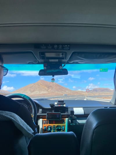 The image captures a view from the driver's seat of a car traveling along a road. The windshield frames a mountainous landscape, with a clear blue sky and some white clouds. The driver is in the foreground, but only the upper portion of their body and the back of their head are visible. They are wearing a dark-colored shirt. The interior of the car is visible, with a black dashboard, various buttons, and a rearview mirror. The rearview mirror is positioned above a small black screen displaying a map. On the passenger side, there is a black rearview mirror and a black phone holder with a smartphone inside. There are also a few knobs and buttons on the passenger side of the dashboard. The coordinates 28.618, -13.856 indicate that the car is located near the village of La Oliva, in the municipality of La Oliva, on the island of Fuerteventura, in the Canary Islands, Spain. The road appears to be a highway, and there is a speed limit sign visible in the distance, which indicates the speed limit is 80 km/h.  The image captures a moment of travel, showcasing the landscape and the interior of the vehicle.