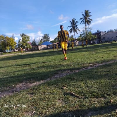 The image shows a soccer field in Nungwi, Tanzania, located at coordinates -5.726, 39.301. The field is covered in short, green grass. There are a few buildings and trees in the background. A young man in a yellow and black striped shirt is running towards the camera, his right leg extended behind him as he walks. There are other players in the background, some of them wearing the same uniform, others in different attire, all playing soccer. There is a "Galaxy A05s" watermark in the lower left-hand corner. The sky is blue with white clouds.