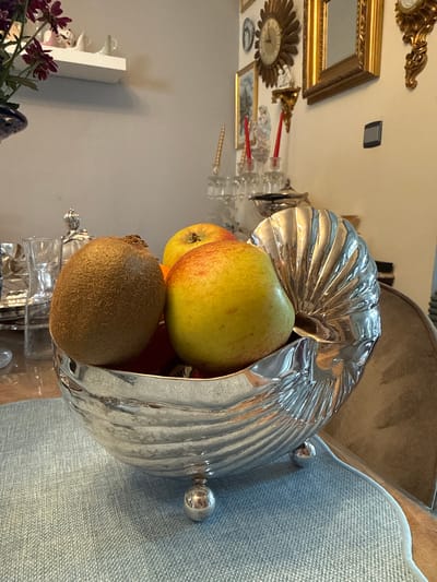 A still life image features a silver, shell-shaped bowl containing fruit. The bowl, which has three spherical feet, rests on a light blue placemat. Inside the bowl are a fuzzy brown kiwi and two apples, one predominantly yellow with red blush and the other mostly red. In the background, a dark blue vase with deep red flowers is partially visible on the left. The scene appears to be indoors, possibly on a dining table, with a wall and decorative items in the background, including framed art, clocks, and candles. The lighting suggests an indoor setting with ambient light sources. The image does not provide any information about the city of Charleroi, Belgium.