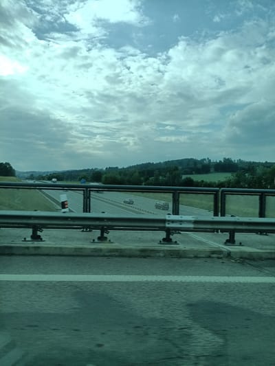 The image depicts a view from an elevated position, likely a bridge or overpass, overlooking a multi-lane highway. The sky is overcast with a mix of light and dark clouds, tinged with a blue-green hue. Below, the highway stretches into the distance, bordered by green fields and dense trees on either side, with rolling hills visible in the background. Several vehicles, including cars and a truck, are on the highway. A metal railing, likely part of the overpass structure, is in the foreground, with concrete barriers and traffic markers visible on the highway below. The image appears to have been taken from a moving vehicle due to motion blur. The location is identified as Šternov, Czechia.