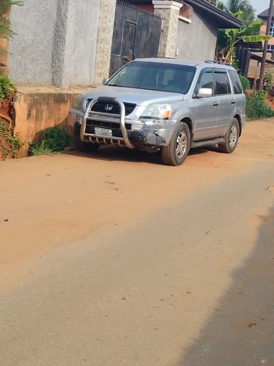 A silver Honda Pilot SUV is parked on the side of a road in Agukwu Nri, Nigeria. The vehicle is positioned facing slightly right, with its front end near a building. It is equipped with a chrome bull bar attached to the front bumper. The front bumper exhibits notable damage and scuffing, particularly on the driver's side, showing black discoloration. A Nigerian license plate, "FST-168AT" from Rivers State, is mounted on the bull bar. The SUV also has roof rails and dark window visors.
The immediate ground beneath the vehicle is an unpaved reddish-brown dirt surface, which transitions into a grey asphalt road in the foreground. Behind the vehicle, a building with a light grey, textured exterior wall and a dark, ornate metal gate is visible. To the left of the vehicle, an exposed reddish-brown earth bank with green vegetation at its base is present. In the background to the right, tropical foliage, including what appears to be banana trees, and a portion of another building with a dark roof are discernible. The scene is illuminated by natural daylight. No individuals are visible.