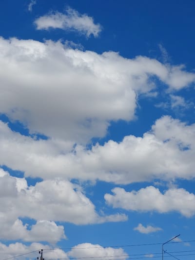 The image shows a clear blue sky with fluffy white cumulus clouds scattered across it. In the lower portion of the image, power lines and a utility pole are visible, suggesting an outdoor urban or suburban environment. The scene is consistent with a bright, sunny day in Kyzylorda, Kazakhstan. There are no people or animals in the frame, and the focus is solely on the sky and the basic infrastructure.