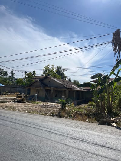 The image shows a rundown wooden house with a damaged roof, located in a somewhat overgrown area. The house appears to be old and in disrepair, with some structural damage visible. The surrounding environment is a mix of vegetation, debris, and a paved road in the foreground. Power lines crisscross the sky above the house and trees. There are no people visible in the image. The scene suggests an abandoned or neglected property, possibly in a rural or semi-urban setting. The location is identified as Bukitrata, Indonesia.