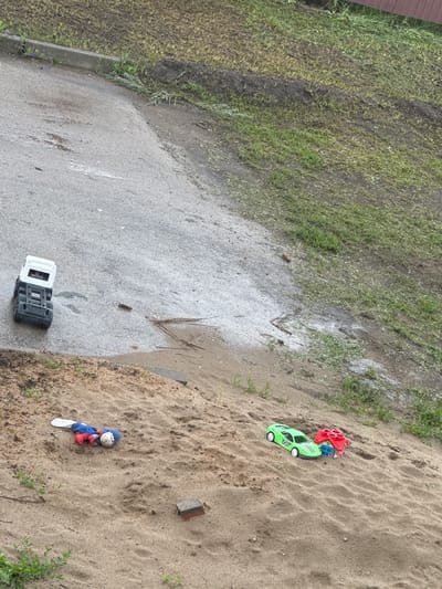 In a sandy area near a paved surface, several toys are scattered. A white and black toy truck is on the paved area, which appears wet in places and has some debris. Further down the sandy slope, a stuffed toy with blue, red, and white elements lies on the sand. Next to the stuffed toy is a small, weathered brick. On the sandy slope, a green toy car and a red toy house are also present. The area is surrounded by grass and dirt. The image is taken from an elevated perspective.