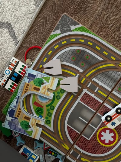 The image displays a wooden toy city play mat on a wooden floor, partially covered by a textured rug. The play mat depicts a city scene with roads, buildings, trees, and traffic signs. A small toy ambulance with red and white stripes and a blue star emblem is positioned on one of the roads, near a building with a red brick facade and a white star symbol within a red circle. Several other toy elements are scattered around, including a small toy house, traffic lights, and road signs. A red string is attached to one of the toy buildings. The scene is lit from above, casting subtle shadows. There are no people visible in the image. The location in the city of Katy, United States is not discernible from the image.